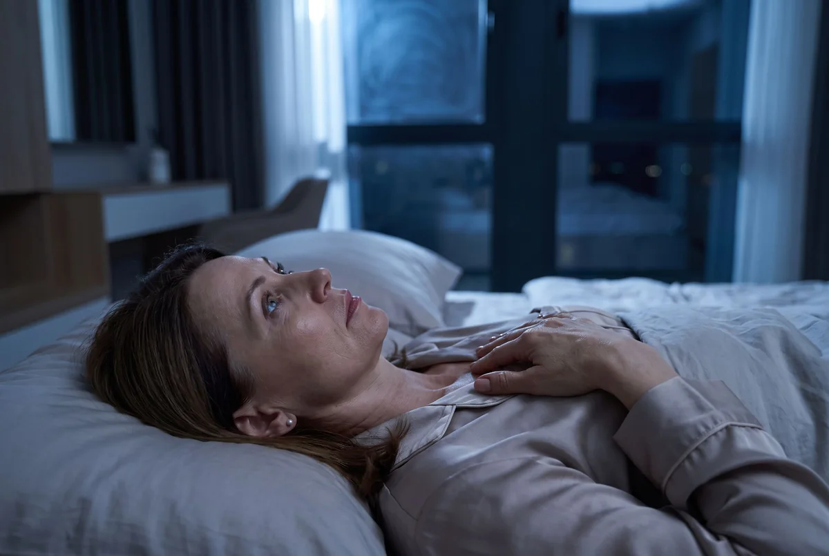 A woman lying awake on her back at night, eyes open, staring at the ceiling, unable to switch off