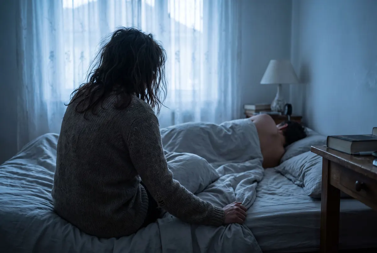 A woman sitting tense on the edge of her bed at dawn while her partner sleeps behind her
